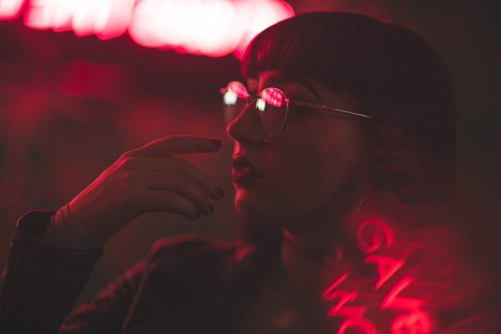 A woman wearing glasses is illuminated by red neon lights in a dimly lit space - Griswold Eye Care A woman wearing glasses is illuminated by red neon lights in a dimly lit space - Griswold Eye Care