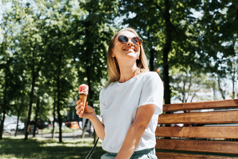 Woman Enjoying Ice Cream on a Sunny Day - Eye Doctor in Norwich, CT Smiling woman in sunglasses enjoys a cone of ice cream while sitting on a bench in a sunlit park surrounded by green trees. - Eye Doctor in Norwich, CT